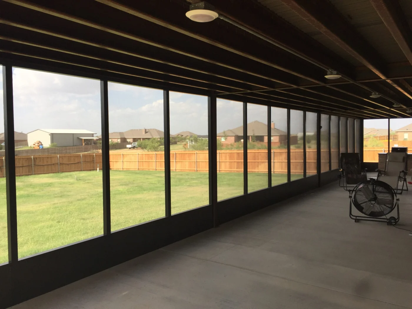 Screened-in patio enclosure with dark frame, concrete floor, ceiling lights, chairs, and a fenced backyard view.
